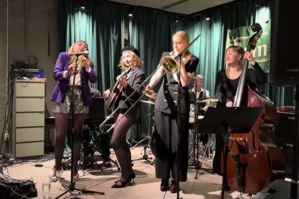Eine Musikkapelle spielt auf der Bühne. Die Musikerinnen treten leidenschaftlich auf und nutzen verschiedene Instrumente.