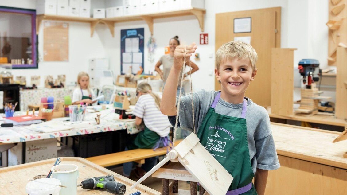 Ein Junge hält stolz ein selbstgebautes Holzprojekt in der Hand. Im Hintergrund arbeiten weitere Kinder an ihren kreativen Projekten. | © www.zloam.at