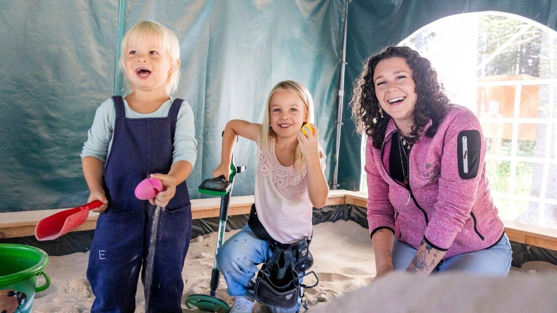 Zwei fröhliche Kinder spielen im Sand mit Schaufeln und einer Schaufel. Eine lächelnde Frau beobachtet sie und genießt den Spaß. | © www.zloam.at