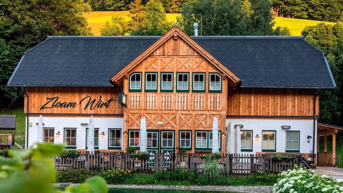 Ein charmantes Holzhaus mit einem traditionellen Design. Umgeben von grünen Bäumen und einem kleinen Teich. | © www.zloam.at
