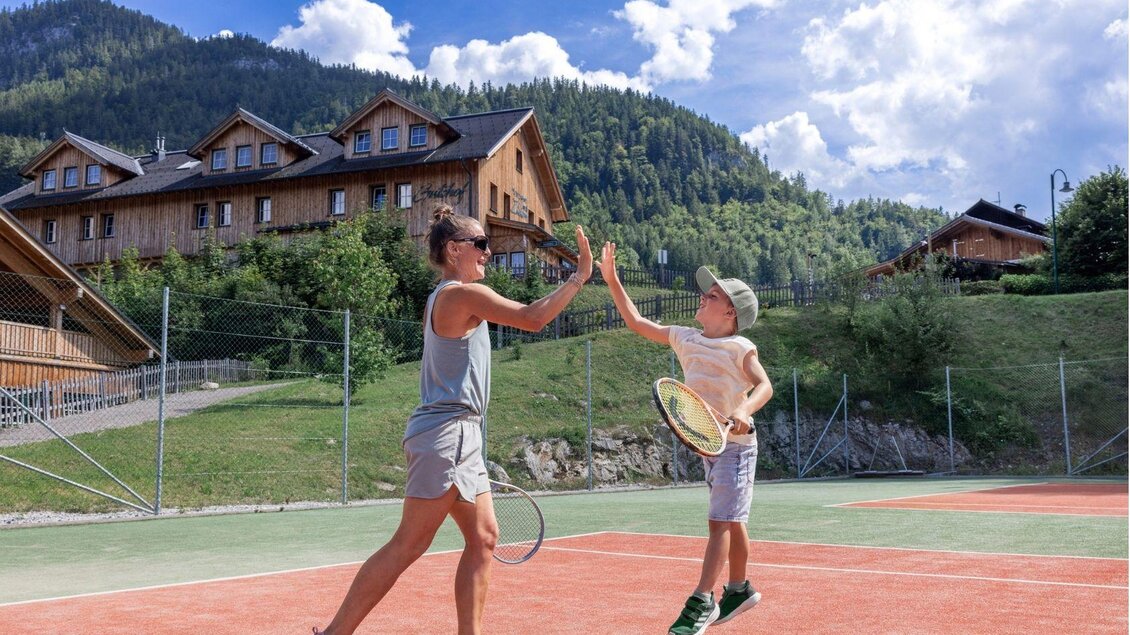 Eine Frau und ein Junge spielen Tennis auf einem Freiplatz. Im Hintergrund sind die Berge und einige Holzgebäude zu sehen. | © www.zloam.at