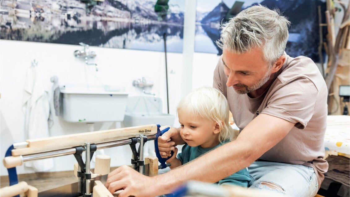 Ein Vater und sein kleines Kind arbeiten gemeinsam an einem Tisch mit Werkzeug. Sie sind in einer hellen Werkstatt umgeben von Holzmaterialien und Werkzeugen. | © www.zloam.at