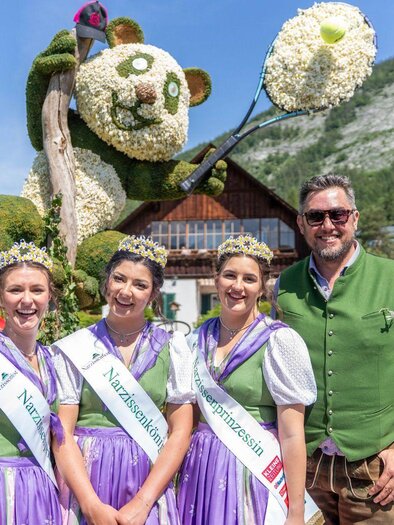 <p>Die drei gekrönten Narzissenhoheiten in traditionellen Trachten mit ihren schönen Kronen von den Narzissenwiesen stehen vor einer Narzissenfigur mit der schönen Landschaft im Hintergrund. | © Narzissenfestverein_Jakob Grill