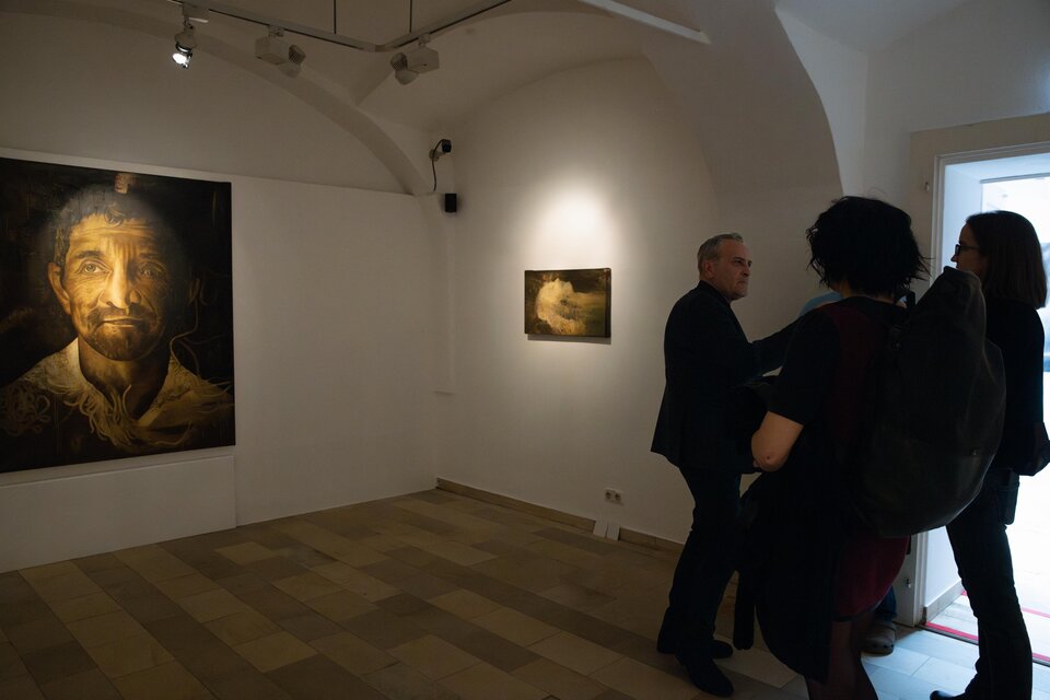 Eine Kunstgalerie mit zwei Bildern an der Wand. Personen stehen zusammen und betrachten die Kunstwerke. | © Achromatic Photography