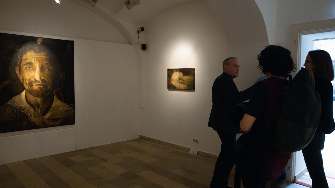 Eine Kunstgalerie mit zwei Bildern an der Wand. Personen stehen zusammen und betrachten die Kunstwerke. | © Achromatic Photography