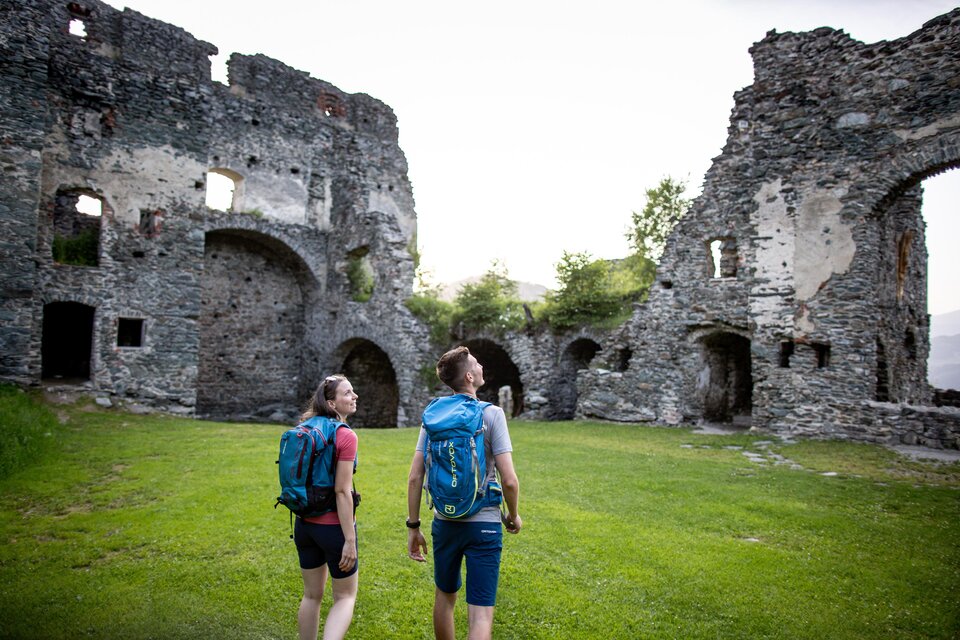 Zwei Wanderer erkunden die Ruinen einer alten Burg. Grünes Gras umgibt die steinernen Mauern, unter einem hellen Himmel. | © Tv Murau