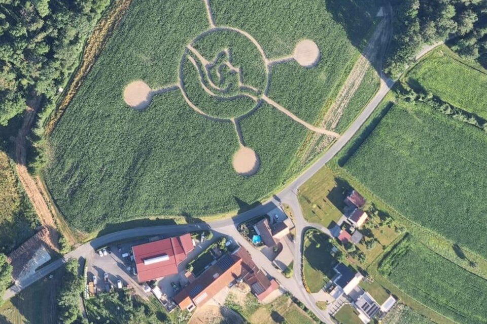 Eine Luftaufnahme eines landwirtschaftlichen Gebiets mit einem auffälligen, geometrischen Muster auf dem Feld. Im Vordergrund sind Häuser und Felder sichtbar, umgeben von Bäumen. | © Lindenhof Kröpfl