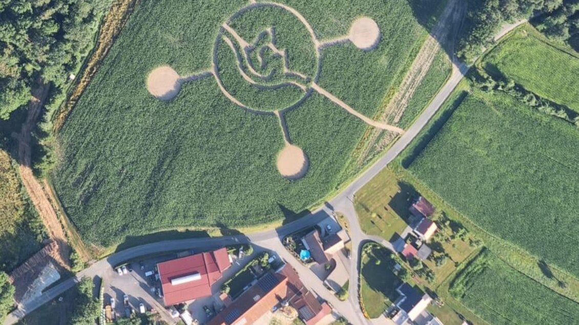Eine Luftaufnahme eines landwirtschaftlichen Gebiets mit einem auffälligen, geometrischen Muster auf dem Feld. Im Vordergrund sind Häuser und Felder sichtbar, umgeben von Bäumen. | © Lindenhof Kröpfl