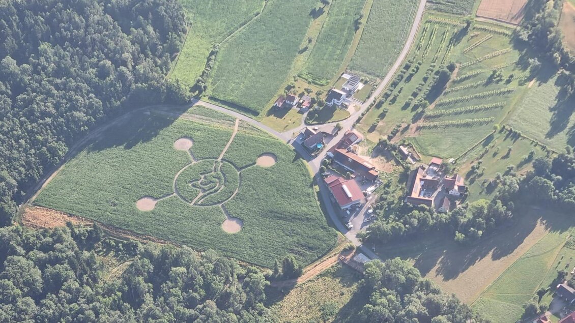 Ein Acker mit einem großen, kreisförmigen Muster und mehreren runden Elementen. In der Nähe sind Häuser und Bäume zu sehen. | © Lindenhof Kröpfl