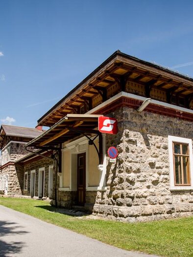 Ein historisches Bahnhofsgebäude mit einer steinernen Fassade. Es liegt in einer ruhigen, naturnahen Umgebung mit Bäumen und Bergen im Hintergrund. | © Bahnhof Neuberg