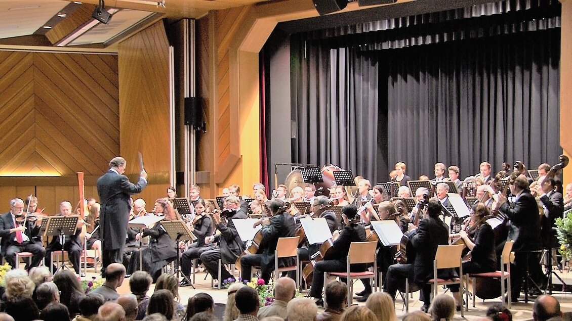 Ein Orchester spielt auf einer Bühne mit einem Dirigenten. Das Publikum sitzt im Vordergrund und hört aufmerksam zu. | © Waltraud Wachmann
