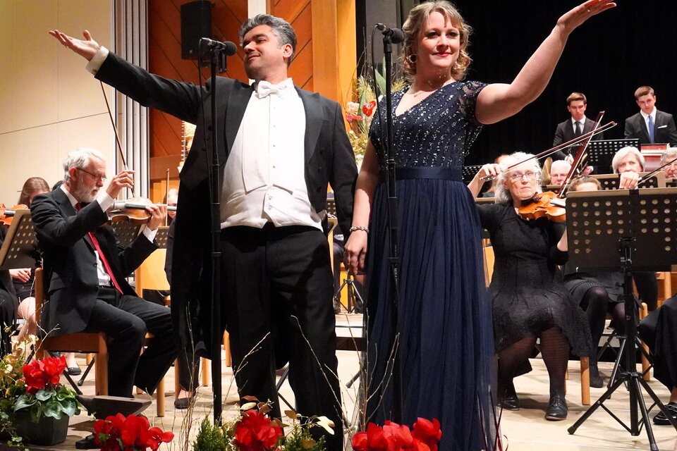 Ein klassisches Konzert mit einem Sängerduo auf der Bühne. Im Hintergrund steht ein Orchester, während Blumenarrangements im Vordergrund zu sehen sind. | © Waltraud Wachmann