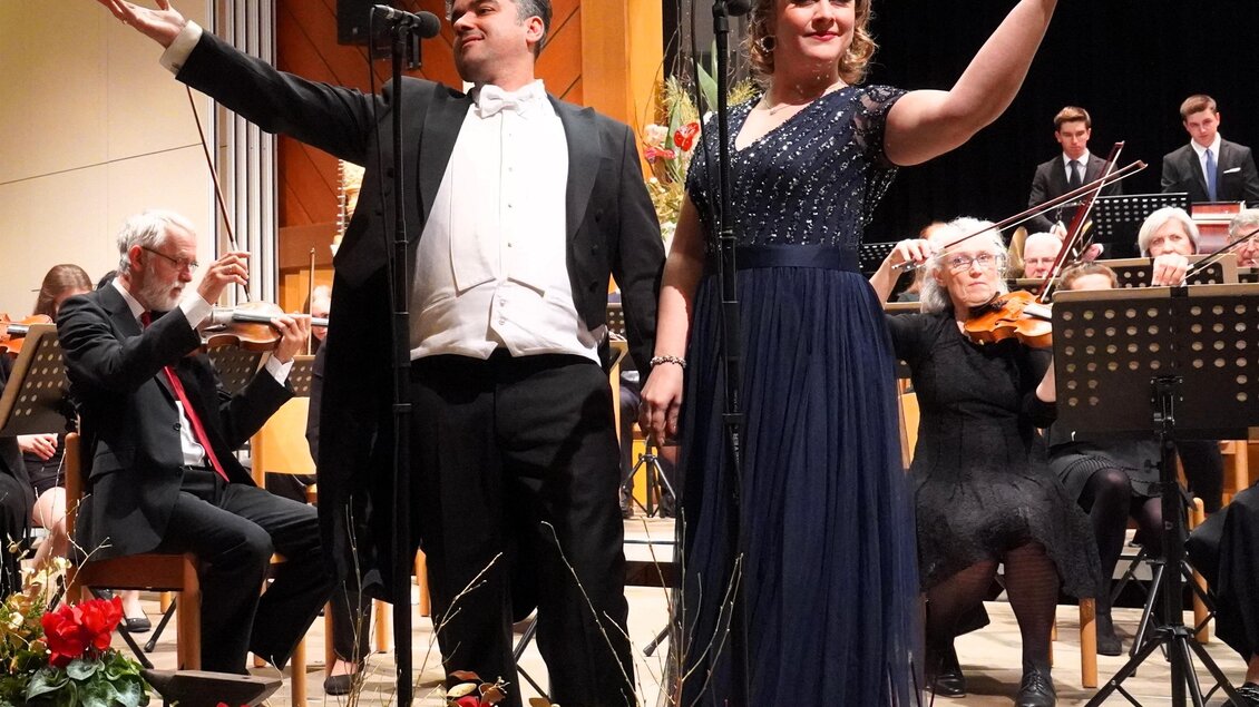 Ein klassisches Konzert mit einem Sängerduo auf der Bühne. Im Hintergrund steht ein Orchester, während Blumenarrangements im Vordergrund zu sehen sind. | © Waltraud Wachmann