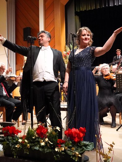 A classical concert with a singer duo on stage. In the background, an orchestra is present, while floral arrangements are visible in the foreground. | © Waltraud Wachmann
