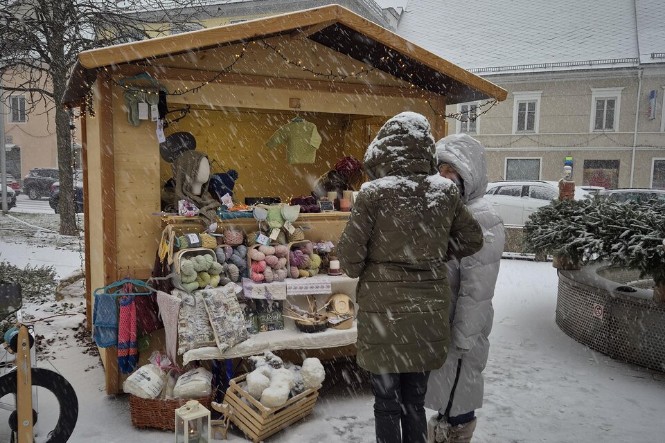 Ein gemütlicher Marktstand im Schnee, umgeben von einer winterlichen Landschaft. Zwei Personen schauen sich die Waren an, während der Schneefall leise fällt. | © TV Murau