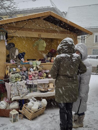 Ein gemütlicher Marktstand im Schnee, umgeben von einer winterlichen Landschaft. Zwei Personen schauen sich die Waren an, während der Schneefall leise fällt. | © TV Murau