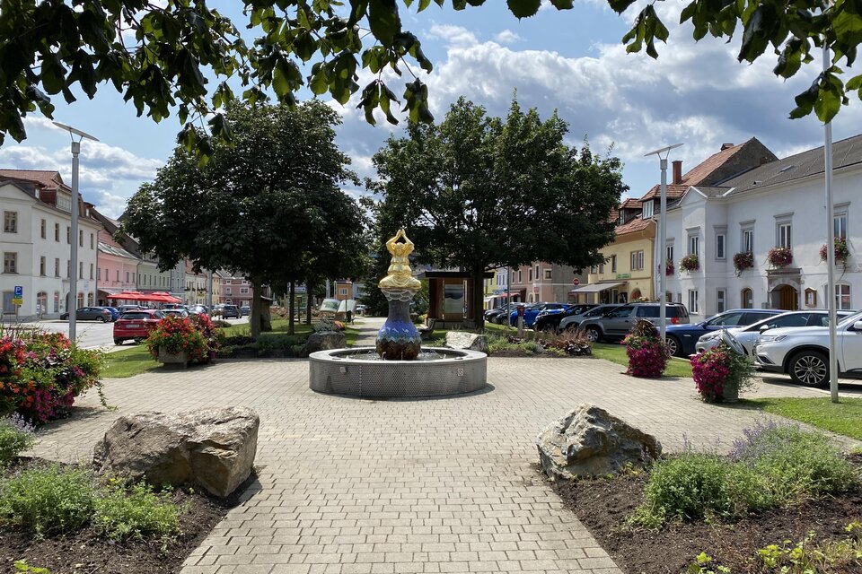 Ein schöner Platz mit einem Brunnen in der Mitte und bunten Blumenbeeten. Umgeben von Bäumen und historischen Gebäuden.