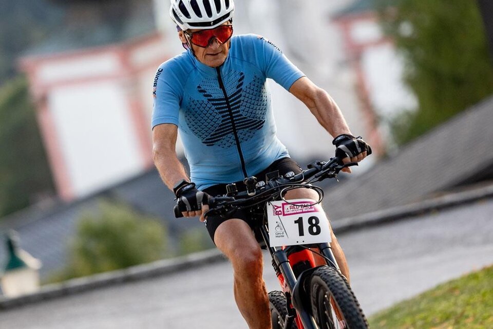 Ein Radfahrer in einem blauen Trikot fährt eine Strecke bergauf. Im Hintergrund sind historische Gebäude und Bäume zu sehen.