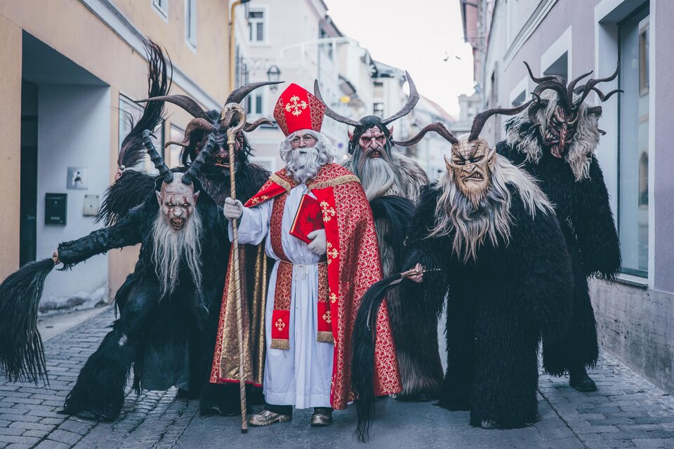 Eine Gruppe von traditionsreich verkleideten Figuren, darunter ein bärtiger Mann in bischöflicher Kleidung und mehrere maskierte Kreaturen. Sie stehen in einer engen Straße und posieren für das Foto. | © Tv Murau