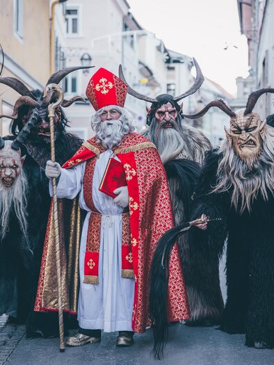 Eine Gruppe von traditionsreich verkleideten Figuren, darunter ein bärtiger Mann in bischöflicher Kleidung und mehrere maskierte Kreaturen. Sie stehen in einer engen Straße und posieren für das Foto. | © Tv Murau