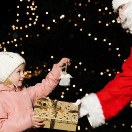 A girl in a pink coat is receiving a gift from a Santa Claus. In the background, many lights are shining. | © Lupi Spuma