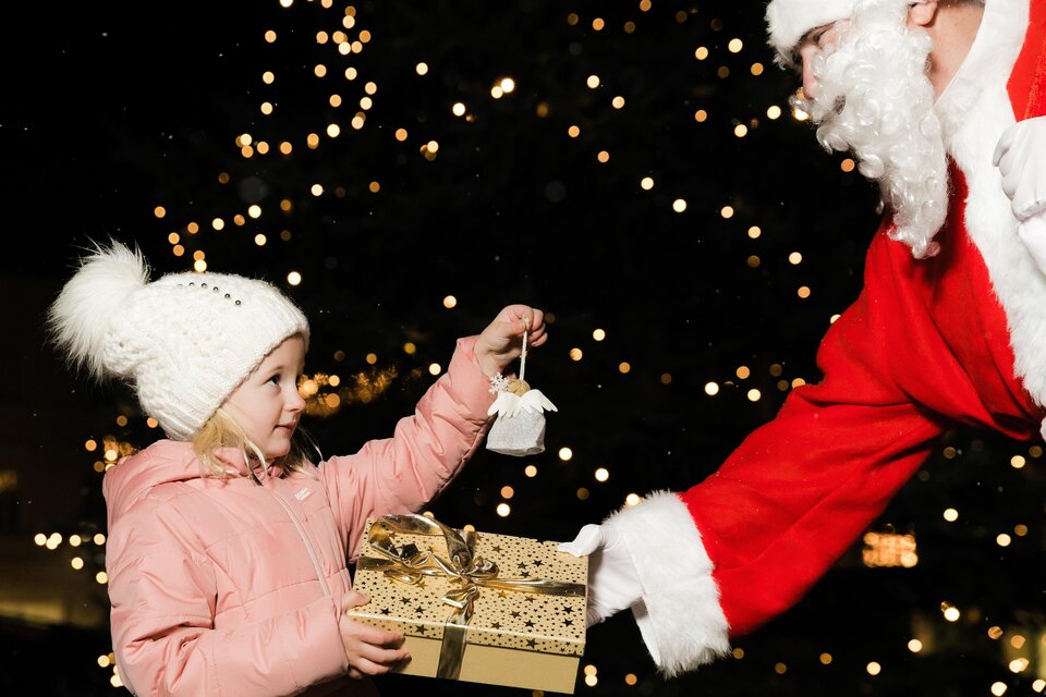 Ein Mädchen in einem rosa Mantel erhält ein Geschenk von einem Weihnachtsmann. Im Hintergrund leuchten viele Lichter. | © Lupi Spuma