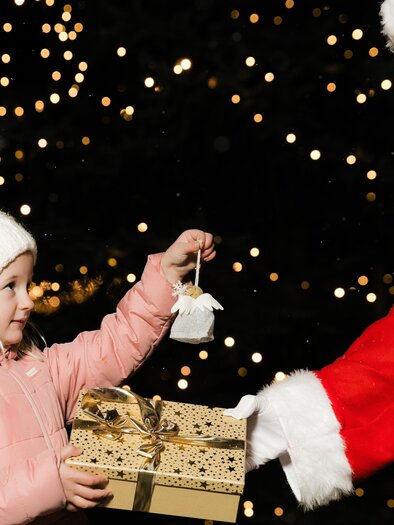 A girl in a pink coat is receiving a gift from a Santa Claus. In the background, many lights are shining. | © Lupi Spuma