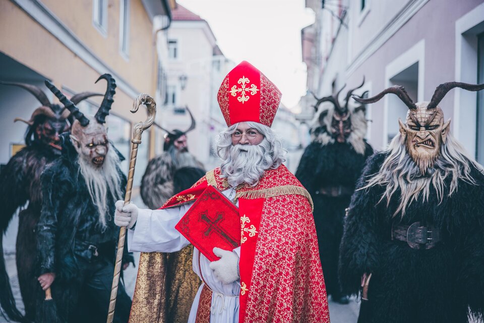 Ein festlich gekleideter Nikolaus mit einem Stab und einem roten Buch steht in einer Straße. Hinter ihm sind mehrere gruselige Figuren mit Hörnern und Fellkostümen. | © TV Murau