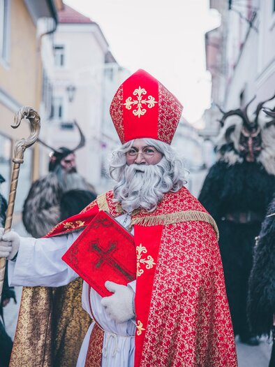 Ein festlich gekleideter Nikolaus mit einem Stab und einem roten Buch steht in einer Straße. Hinter ihm sind mehrere gruselige Figuren mit Hörnern und Fellkostümen. | © TV Murau