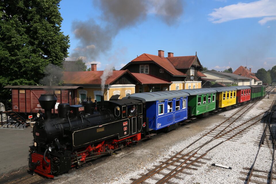 Eine dampfbetriebene Lokomotive mit bunten Waggons steht an einem Bahnhof. Im Hintergrund sind Bäume und ein blauer Himmel zu sehen. | © TV Südsteiermark - Dominik Feibel