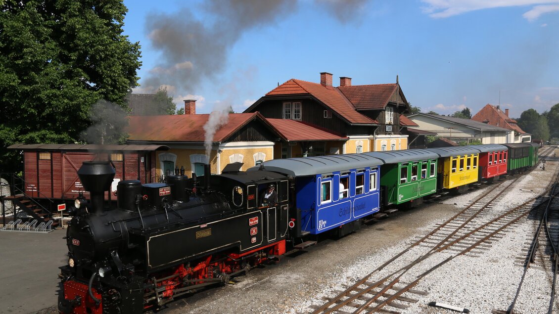 Eine dampfbetriebene Lokomotive mit bunten Waggons steht an einem Bahnhof. Im Hintergrund sind Bäume und ein blauer Himmel zu sehen. | © TV Südsteiermark - Dominik Feibel