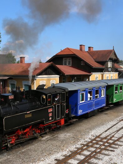Eine dampfbetriebene Lokomotive mit bunten Waggons steht an einem Bahnhof. Im Hintergrund sind Bäume und ein blauer Himmel zu sehen. | © TV Südsteiermark - Dominik Feibel