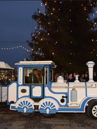 A festive little train stands in front of a large, decorated Christmas tree. The surroundings are illuminated in the evening, and people are standing nearby. | © Stadtgemeinde Deutschlandsberg