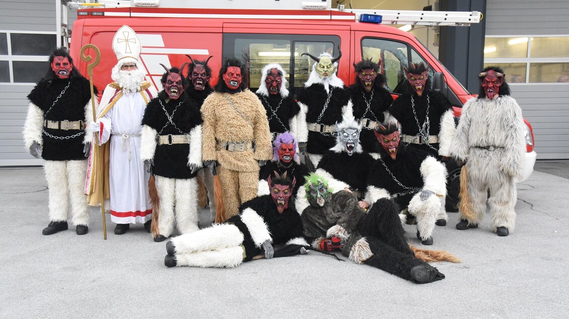 Eine Gruppe von Personen in festlichen Kostümen, darunter Figuren mit roten Gesichtern und traditionellen Kleidern, posiert vor einem Feuerwehrfahrzeug. Die Szene zeigt eine kulturelle Feier oder Veranstaltung. | © Tourimusverband Oststeiermark