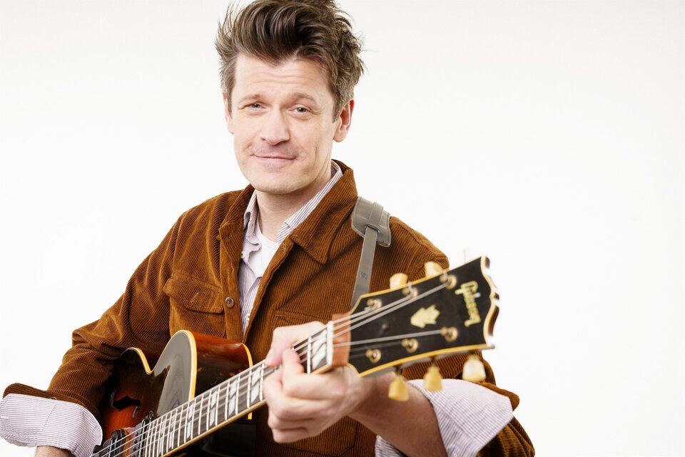 Norbert Schneider trägt eine leichte Braune Jacke über einem Hemd und hält seine Gitarre im Arm. | © Tourismusverband Oststeiermark