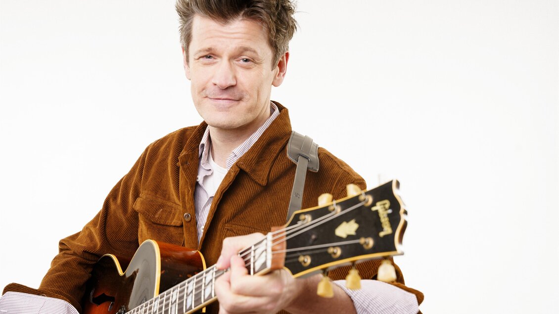 Norbert Schneider trägt eine leichte Braune Jacke über einem Hemd und hält seine Gitarre im Arm. | © Tourismusverband Oststeiermark