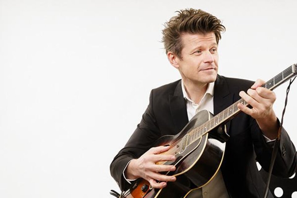 Ein Mann in einem Anzug spielt Gitarre und lächelt. Der Hintergrund ist schlicht und hell. | © Georg Buxhofer