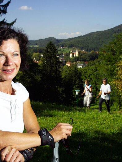 Eine Frau steht in einer grünen Landschaft mit Bergen im Hintergrund. Im Hintergrund sind weitere Personen zu sehen, die die Natur genießen. | © TVB Thermen- & Vulkanland