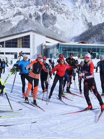 Eine Gruppe von Skifahrern steht auf einer verschneiten Fläche und bereitet sich auf den Langlauf vor. Im Hintergrund sind schneebedeckte Berge und ein modernes Gebäude zu sehen. | © Michael Simonlehner