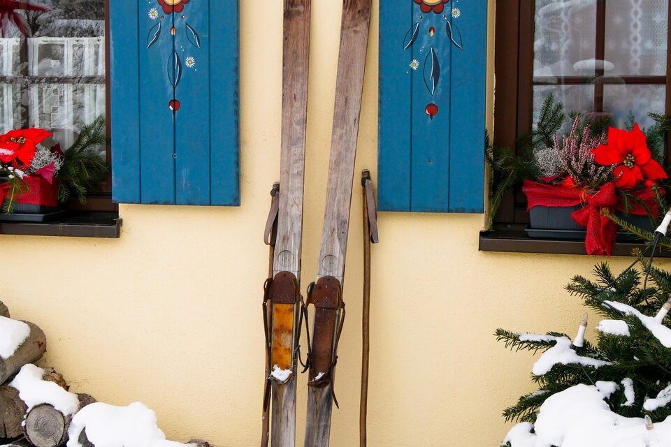 Zwei Holzskier stehen an einer Wand mit blauen Fensterläden. Im Vordergrund liegt Schnee und einige Holzscheite sind gestapelt. | © Pixabay