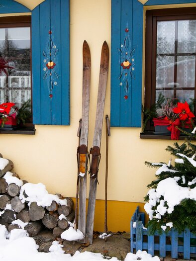 Zwei Holzskier stehen an einer Wand mit blauen Fensterläden. Im Vordergrund liegt Schnee und einige Holzscheite sind gestapelt. | © Pixabay