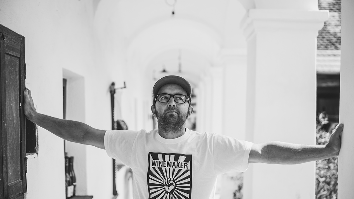 Die Schwarz-Weiß-Aufnahme zeigt einen Winzer mit Brille, Bart und Kappe in einem historischen Arkadengang. Mit weit ausgestreckten Armen lehnt er sich an die Wände und blickt nachdenklich zur Seite. Sein T-Shirt mit der Aufschrift „Winemaker Independent“ unterstreicht seine Leidenschaft für unabhängigen Weinbau. Das Foto verbindet Tradition, Persönlichkeit und moderne Weinkultur. | © Bernhard Bergmann