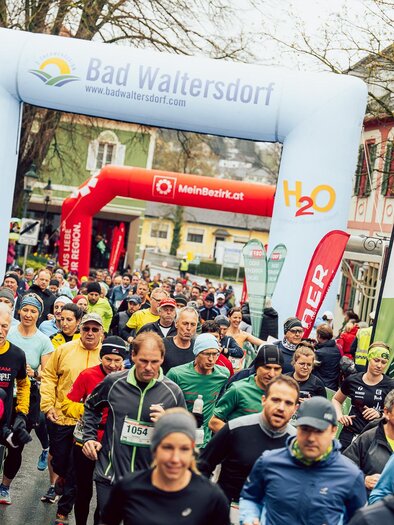 Eine Gruppe von Läufern startet bei einem Rennen in Bad Waltersdorf. Bunte Sportbekleidung und ein großer Startbogen prägen die Szene. | © Hikimus