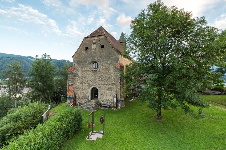 Ein historisches Gebäude aus Stein, umgeben von grüner Wiese und Bäumen. Im Hintergrund sind sanfte Hügel und ein klarer Himmel zu sehen. | © fotodesignbyMarcus Auer