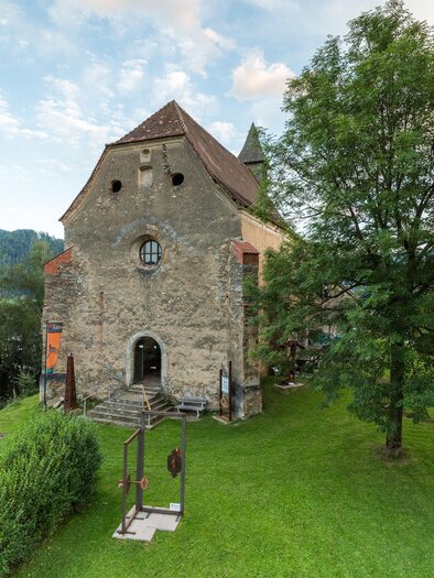 Ein historisches Gebäude aus Stein, umgeben von grüner Wiese und Bäumen. Im Hintergrund sind sanfte Hügel und ein klarer Himmel zu sehen. | © fotodesignbyMarcus Auer