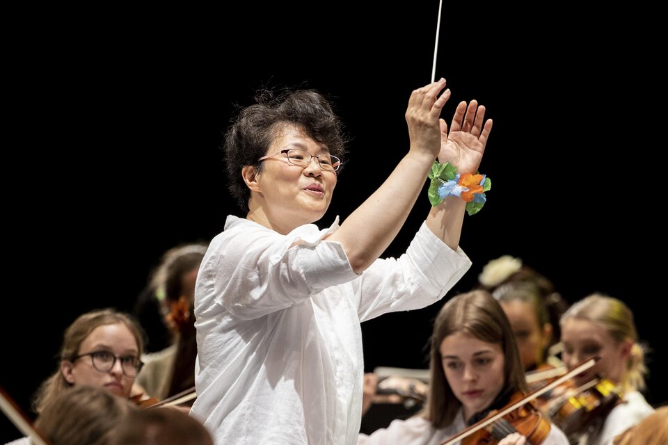 Eine Dirigentin leitet ein Orchester und hebt ihren Stock. Im Hintergrund spielen junge Musikerinnen Geige. | © Nikola Milatovic