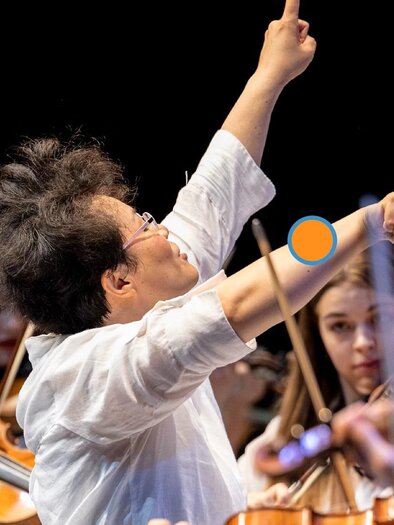 Ein Dirigent steht auf der Bühne und leitet ein Orchester. Im Hintergrund sind Musiker mit ihren Instrumenten zu sehen. | © Nikola Milatovic
