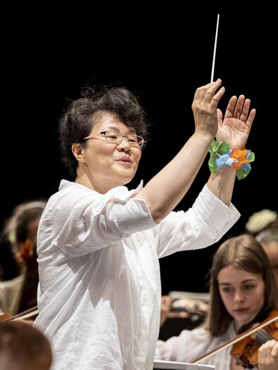 A conductor leads an orchestra and raises her baton. In the background, young female musicians are playing the violin. | © Nikola Milatovic