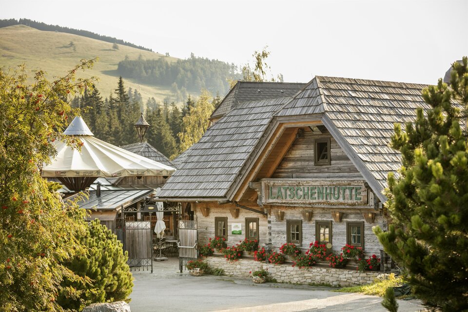 Eine gemütliche Alm in den Bergen mit Holzfassade und blühenden Fenstern. Umgeben von grünen Hügeln und Bäumen. | © Latschenhütte