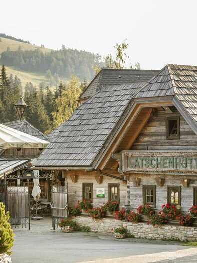 Egy kényelmes alpesi ház a hegyekben, fából készült homlokzattal és virágzó ablakokkal. Zöld dombokkal és fákkal körülvéve. | © Latschenhütte